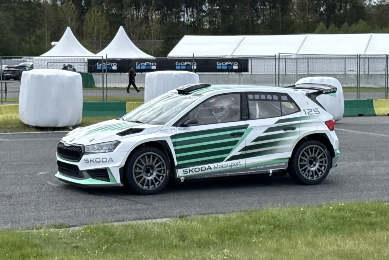 Samochód rajdowy Škoda Fabia z charakterystycznym zielono-białym malowaniem i napisem Skoda Motorsport, stojący na torze wyścigowym, otoczony elementami zabezpieczającymi oraz ogrodzeniem w tle.