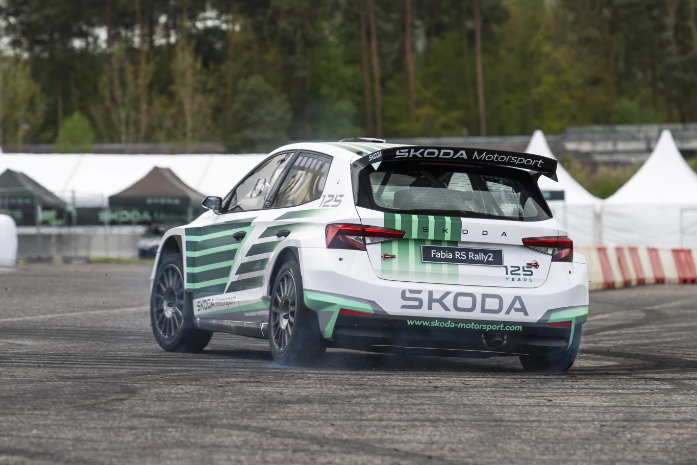Samochód rajdowy Škoda Fabia RS Rally2 w charakterystycznych biało-zielonych barwach pokonuje zakręt na torze asfaltowym, za nim widoczne są bandy ochronne i namioty serwisowe.