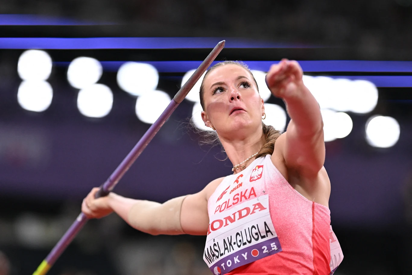Małgorzata Maślak-Glugla Zawodniczka w biało-czerwonym stroju sportowym z napisem POLSKA koncentruje się podczas rzutu oszczepem na lekkoatletycznych zawodach, trzymając oszczep w pozycji do dynamicznego wyrzutu.