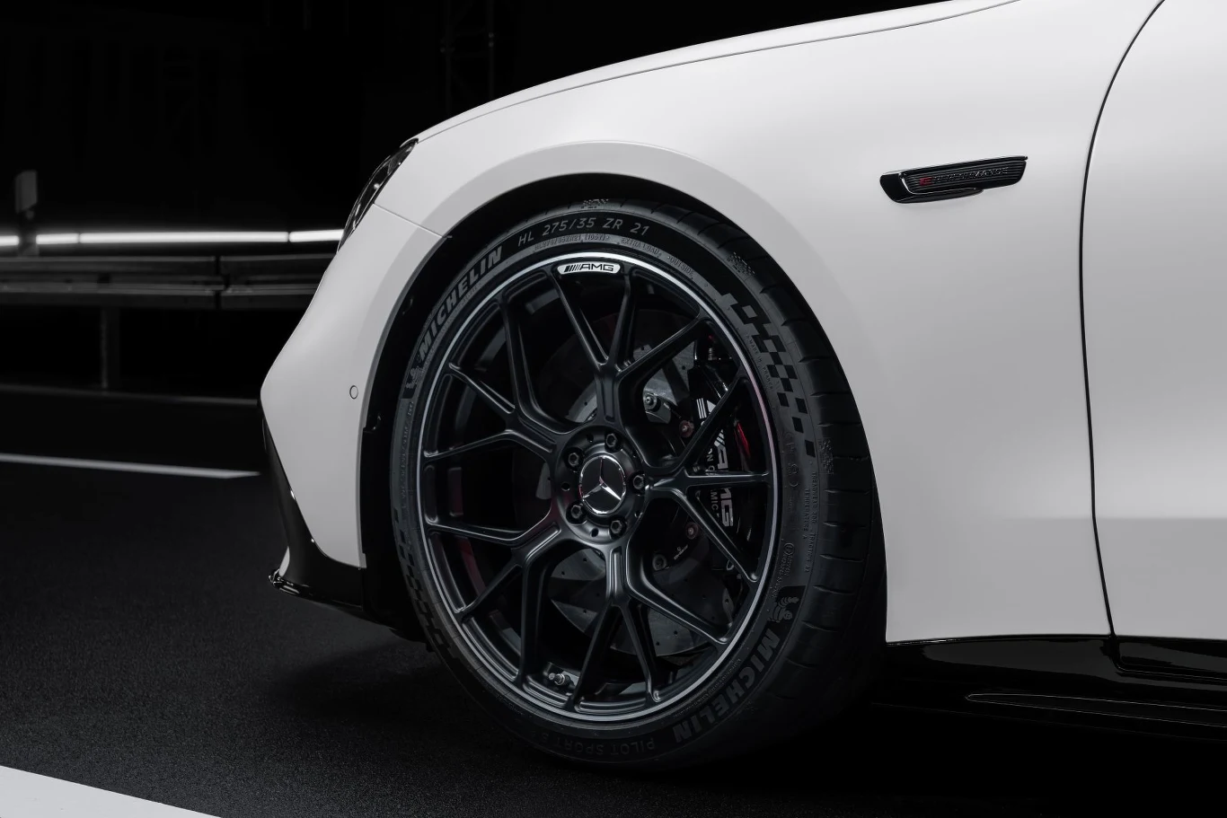 Czarny, sportowy felga z logo Mercedes-AMG na tle białego nadwozia samochodu, wyeksponowane przednie koło z widocznym zaciskiem hamulcowym.