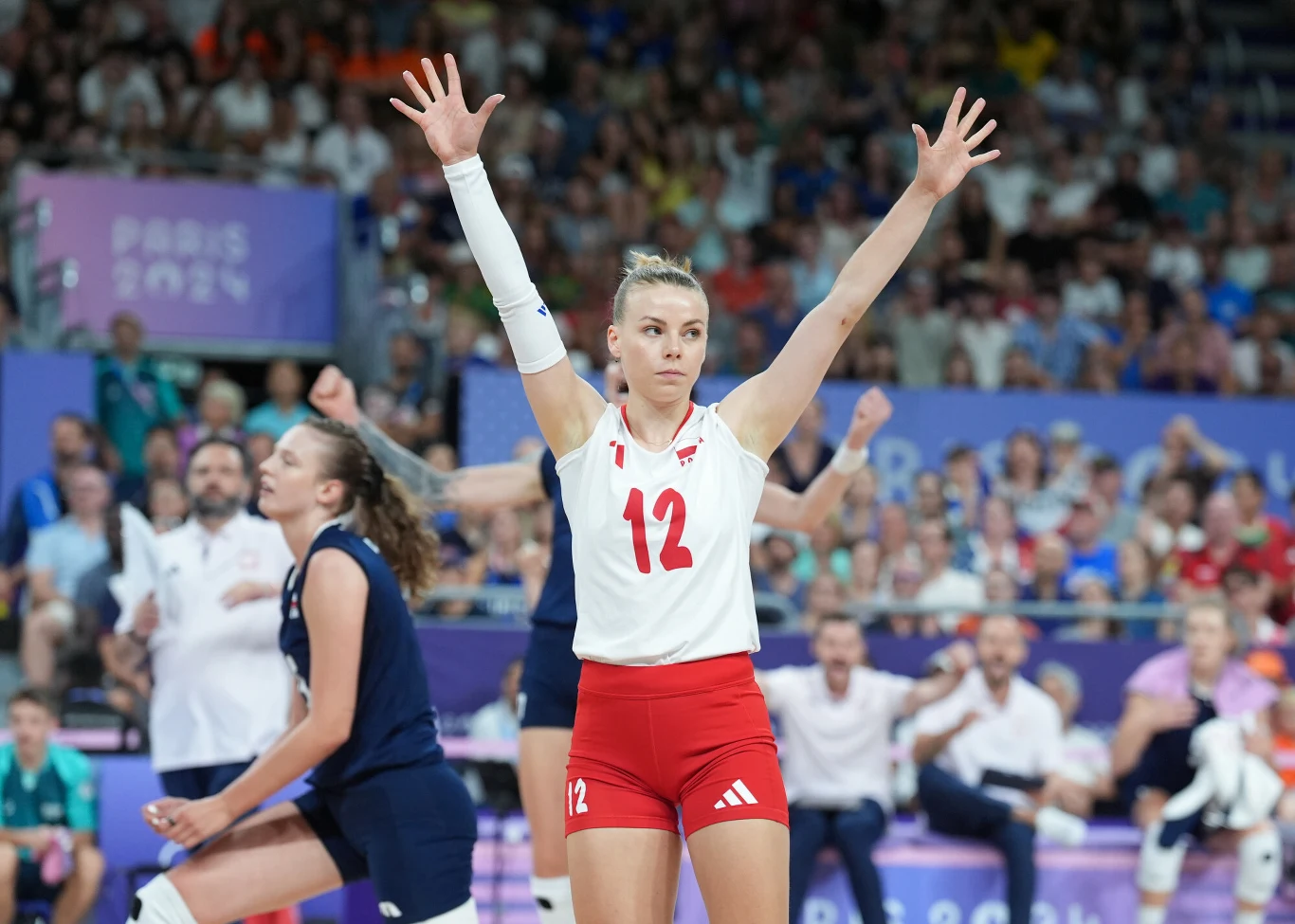 Siatkarka w białej koszulce z numerem 12 i czerwonych spodenkach, stojąca na boisku z uniesionymi rękami, otoczona przez inne zawodniczki w trakcie meczu na hali sportowej.
