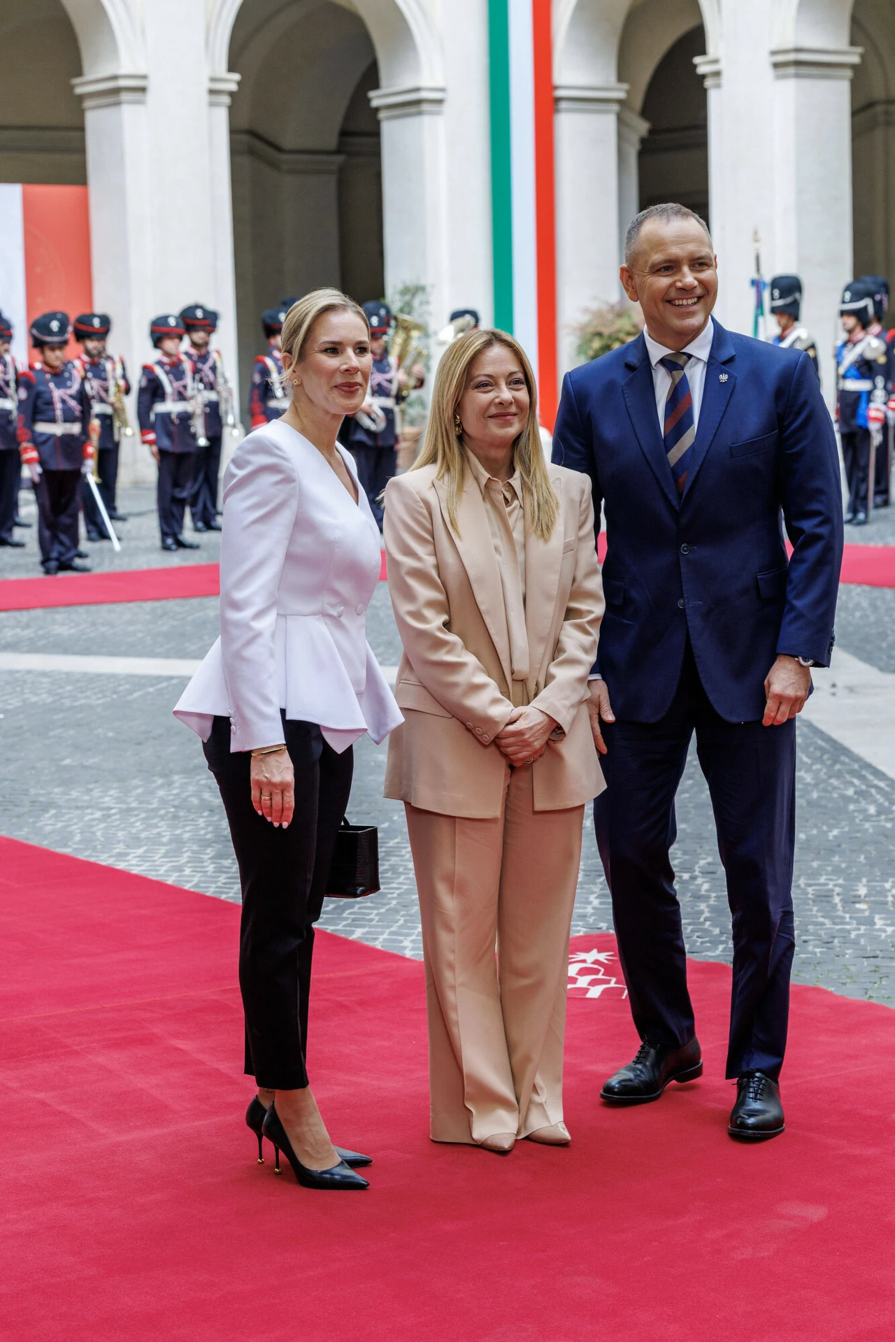 Trzy elegancko ubrane osoby stoją na czerwonym dywanie, w tle żołnierze w uroczystych mundurach oraz duża pionowa flaga w barwach włoskich.