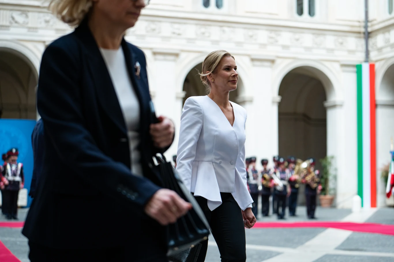 Kobieta w białej marynarce idzie po czerwonym dywanie w otoczeniu elegancko ubranych osób, w tle widoczna orkiestra wojskowa oraz pionowa włoska flaga przy kolumnach pałacu.