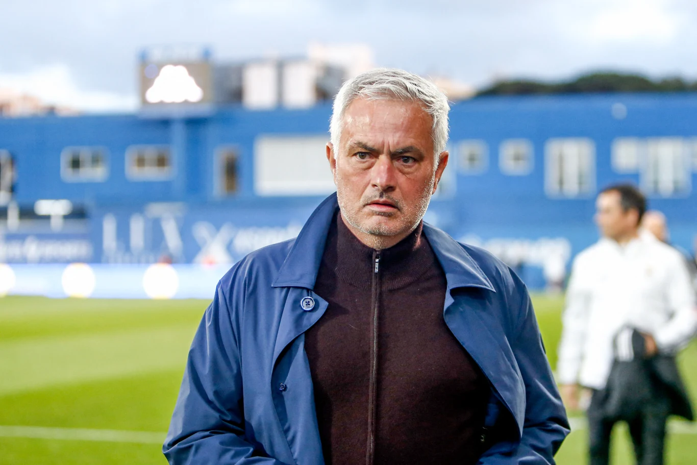 Jose Mourinho Mężczyzna o siwych włosach w ciemnej kurtce stoi na tle niebieskiego budynku przy boisku sportowym, z poważnym wyrazem twarzy.
