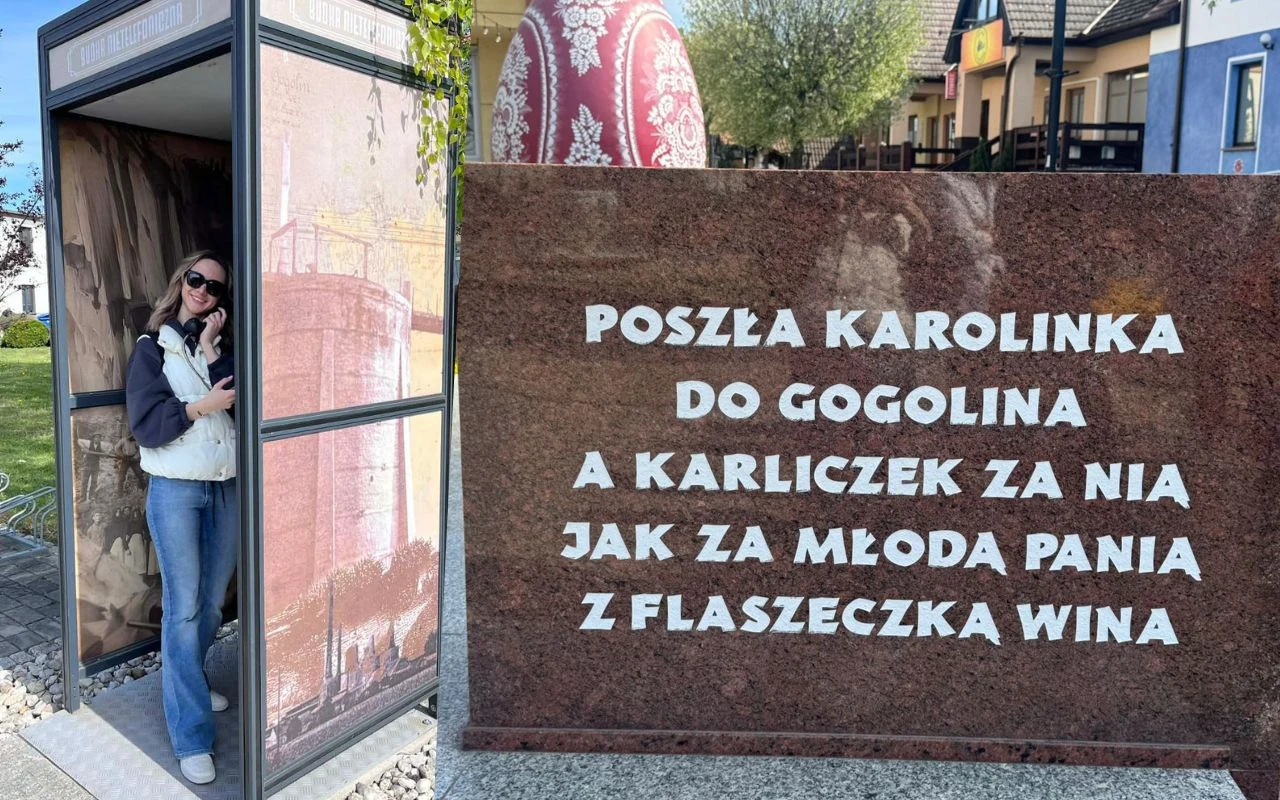Kobieta w okularach przeciwsłonecznych stoi w starej, oszklonej budce telefonicznej, obok której znajduje się kamienna tablica z wyrytym tekstem popularnej śląskiej piosenki ludowej.