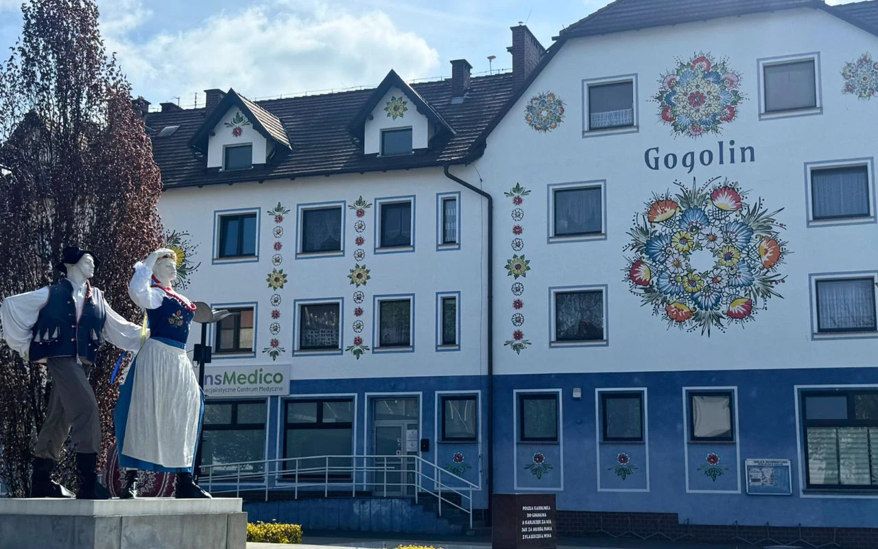 Kolorowy mural z kwiatowymi motywami zdobi ścianę budynku w Gogolinie, przed którym stoi pomnik pary ubranej w tradycyjne śląskie stroje ludowe.