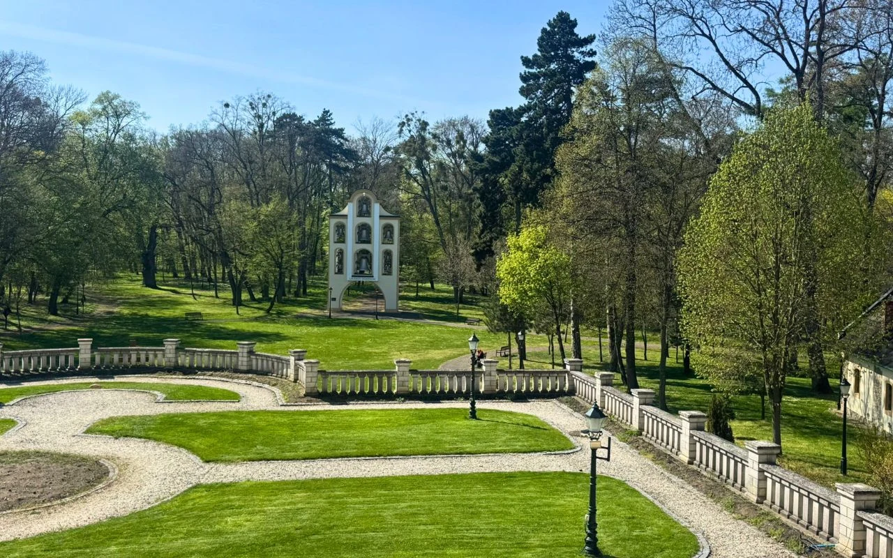 Zadbany park z alejkami wysypanymi żwirem, ozdobną kamienną balustradą i charakterystyczną białą budowlą przypominającą dzwonnicę lub kapliczkę pośród drzew.