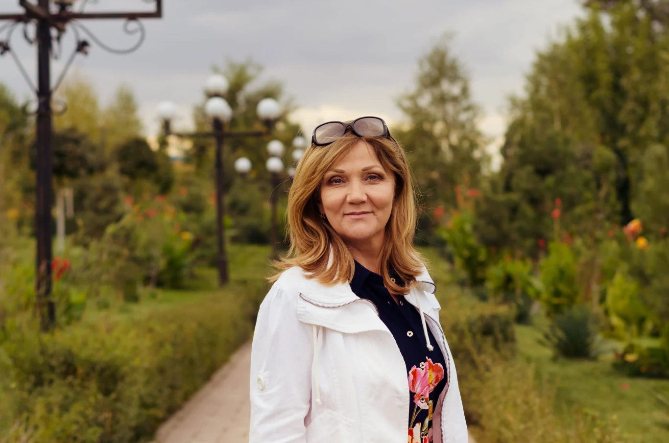 Kobieta z blond włosami i okularami na głowie stoi na alejce otoczonej zielenią i kwiatami, w tle stylowe latarnie i drzewa.