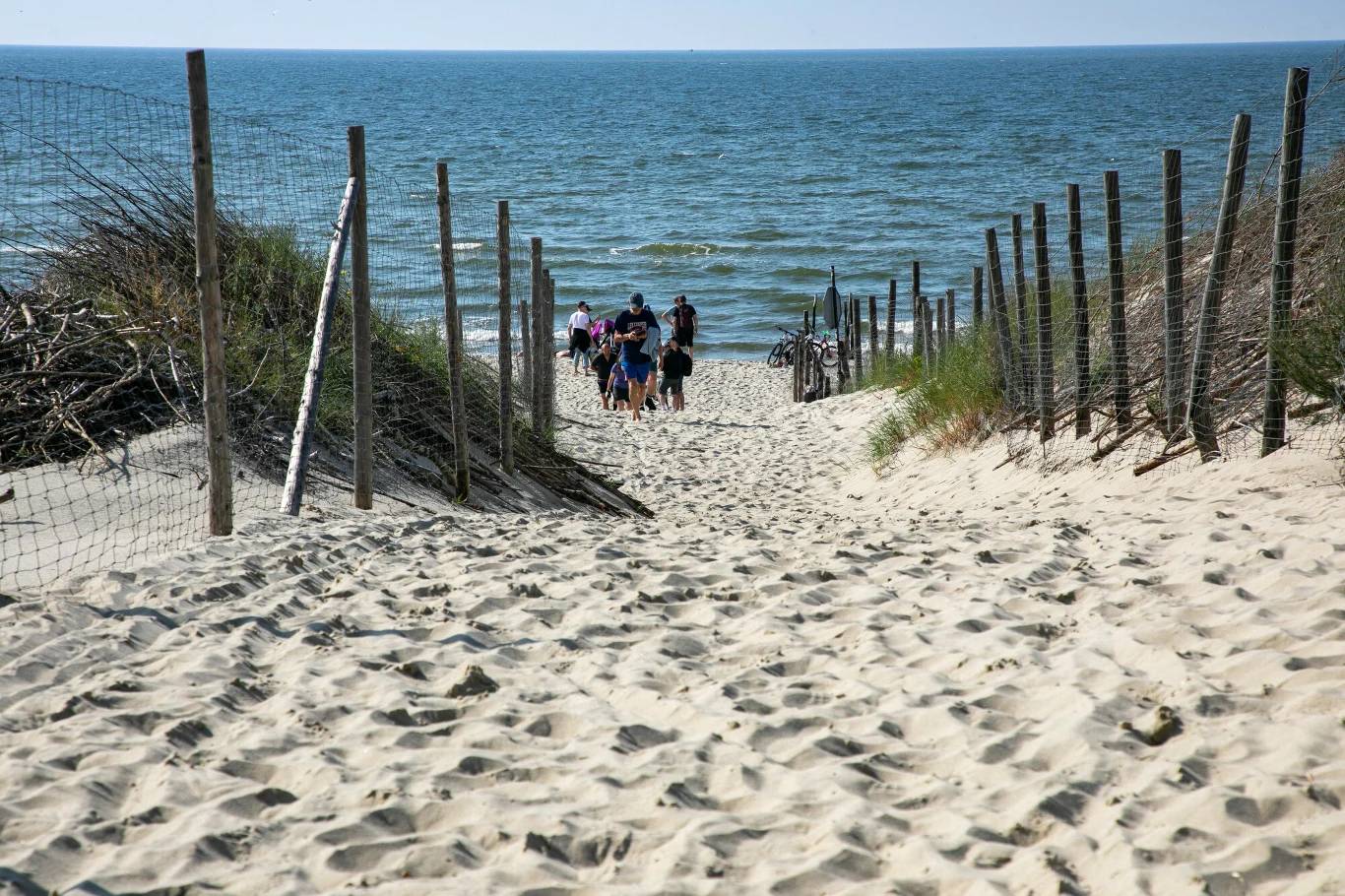 Plaża w Piaskach Szeroka, piaszczysta ścieżka wśród wydm prowadząca do morza, grupka osób idąca w stronę wody, po bokach płoty z siatki i trawy porastające piasek.