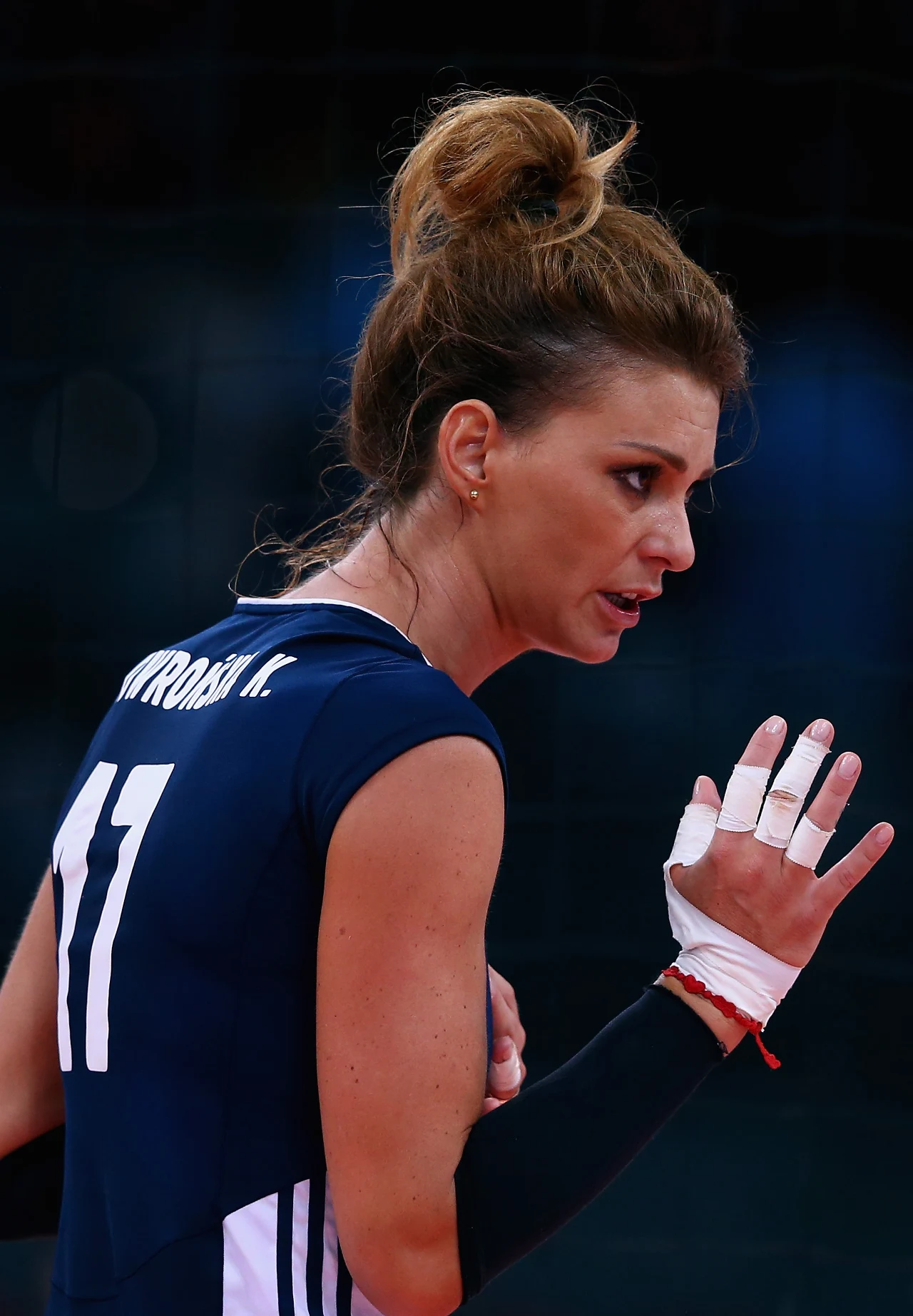 Siatkarka w granatowym stroju sportowym z numerem 11, z włosami upiętymi w luźny kok i zabandażowaną dłonią uniesioną w geście komunikacji podczas meczu.
