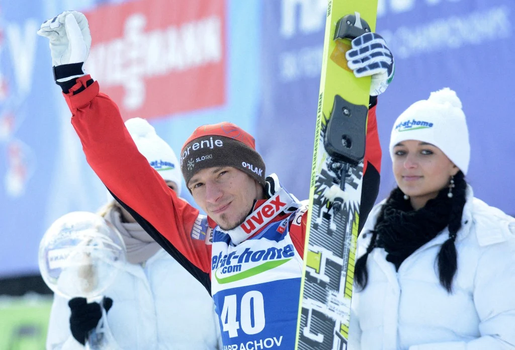 Skoczek narciarski w kombinezonie startowym, trzymający narty pionowo i unoszący rękę w geście triumfu, w otoczeniu osób ubranych w zimowe stroje i czapki z logo sponsora.