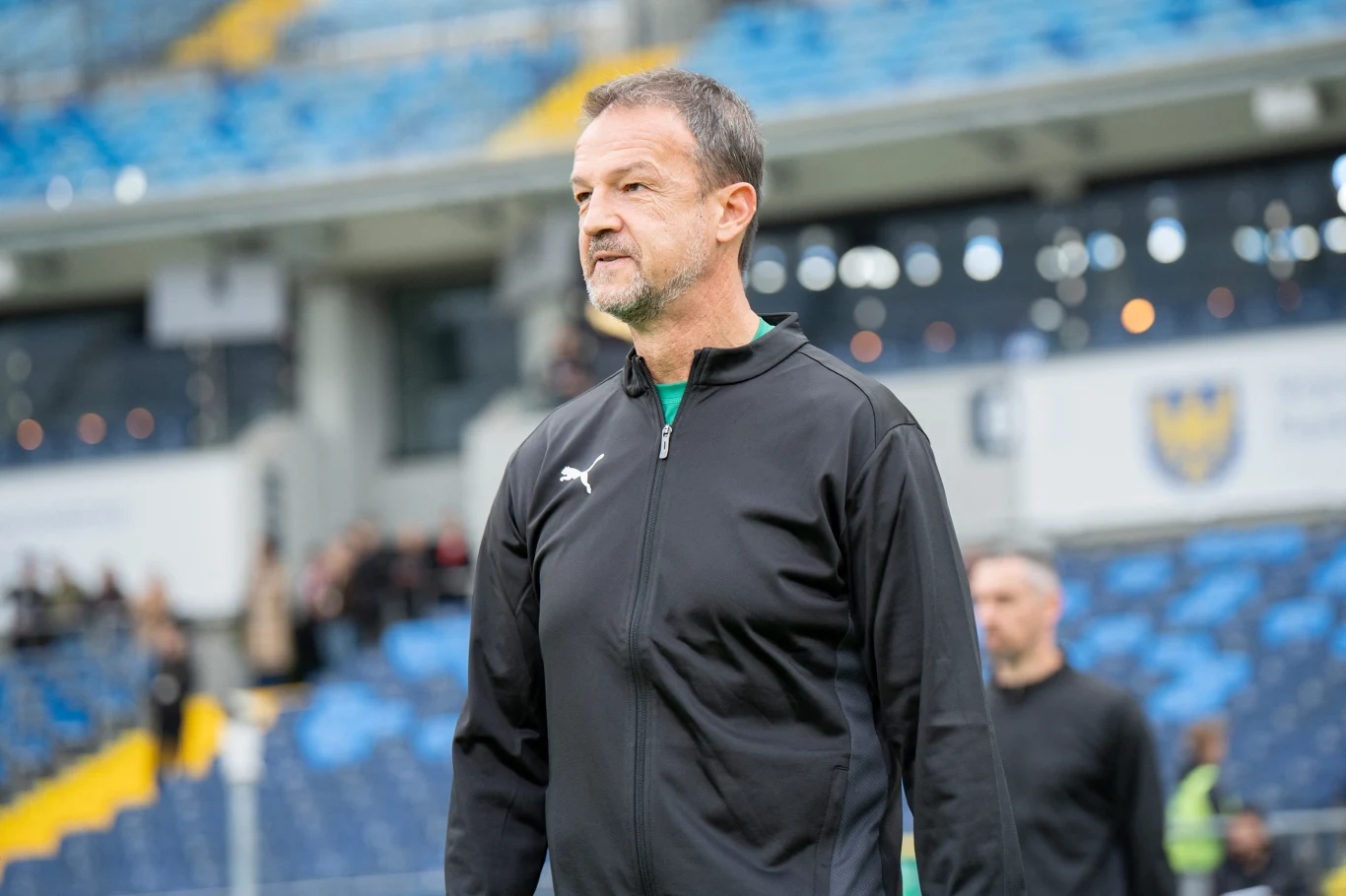 Fredy Bobic Mężczyzna w czarnej sportowej kurtce stojący na tle pustych niebieskich trybun stadionu.