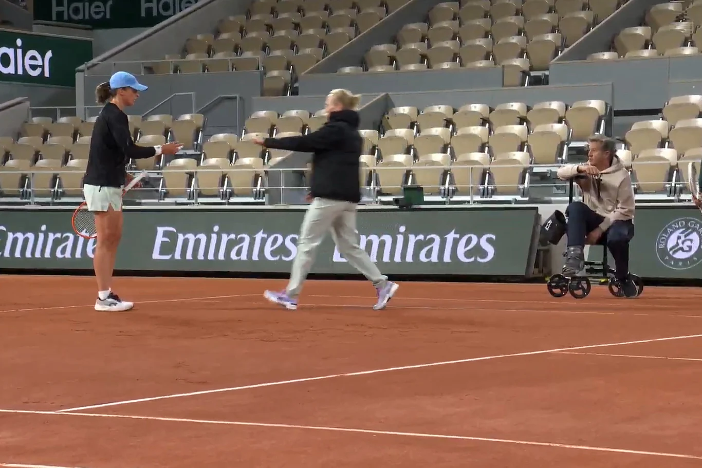 Dwie kobiety spotykają się na korcie tenisowym, jedna ubrana sportowo trzyma rakietę, druga biegnie w jej kierunku z rozłożonymi ramionami, w tle widoczna osoba siedząca na chodziku oraz puste trybuny stadionu.