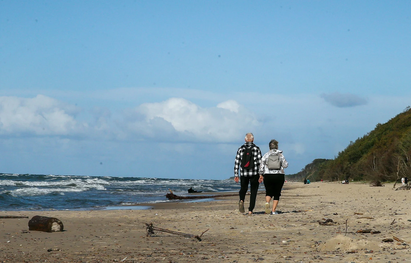 Dwie osoby spacerujące brzegiem morza w kierunku oddalonego klifu, piaszczysta plaża z rozrzuconymi fragmentami drewna, spokojne fale i jasne niebo z pojedynczymi chmurami.