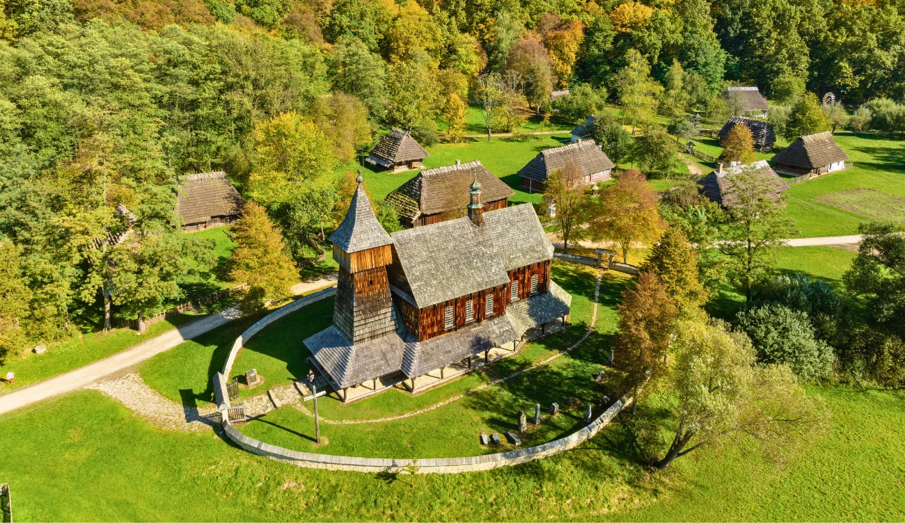 Drewniany kościół otoczony kamiennym murkiem, położony na zielonej polanie, w sąsiedztwie kilku tradycyjnych drewnianych budynków i gęstego lasu w tle.