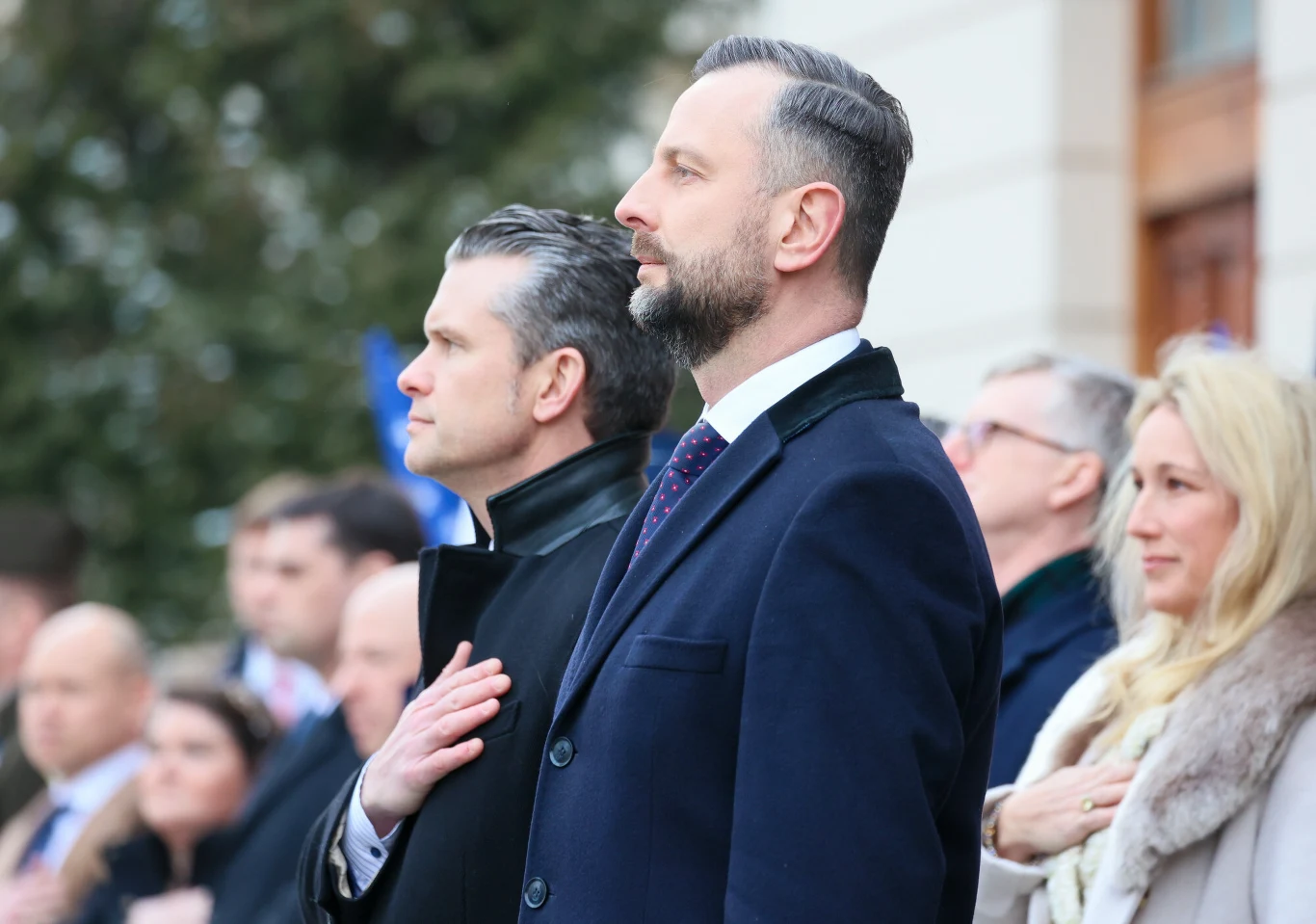 Pete Hegseth, sekretarz obrony Stanów Zjednoczonych, oraz Władysław Kosiniak-Kamysz, wicepremier i szef MON Grupa elegancko ubranych osób stoi w skupieniu, z rękami położonymi na sercu, zwrócona twarzą w jednym kierunku.