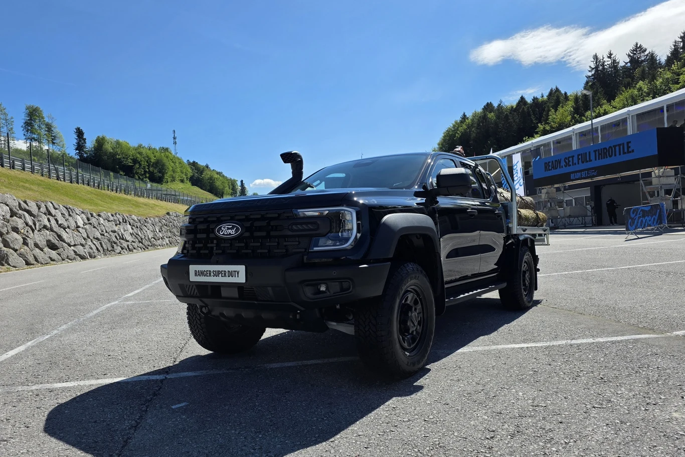 Czarny, masywny pickup Ford z podwyższonym zawieszeniem i akcesoriami terenowymi ustawiony na asfaltowym placu przy torze wyścigowym, w otoczeniu zielonych wzgórz i nowoczesnej zabudowy.