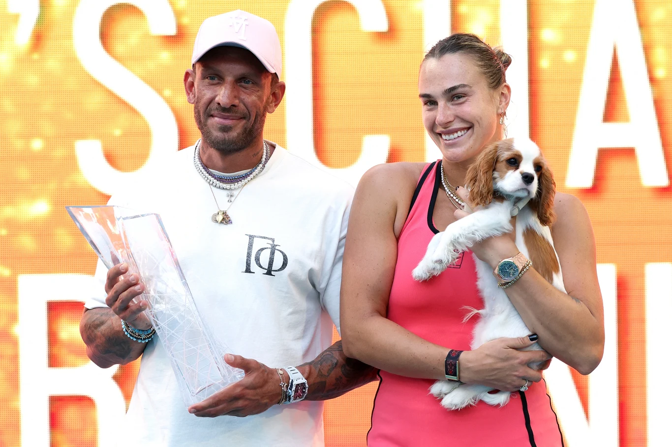 Mężczyzna trzymający przezroczyste trofeum oraz uśmiechnięta kobieta w sportowym stroju obejmująca małego psa; w tle duży, rozświetlony napis.