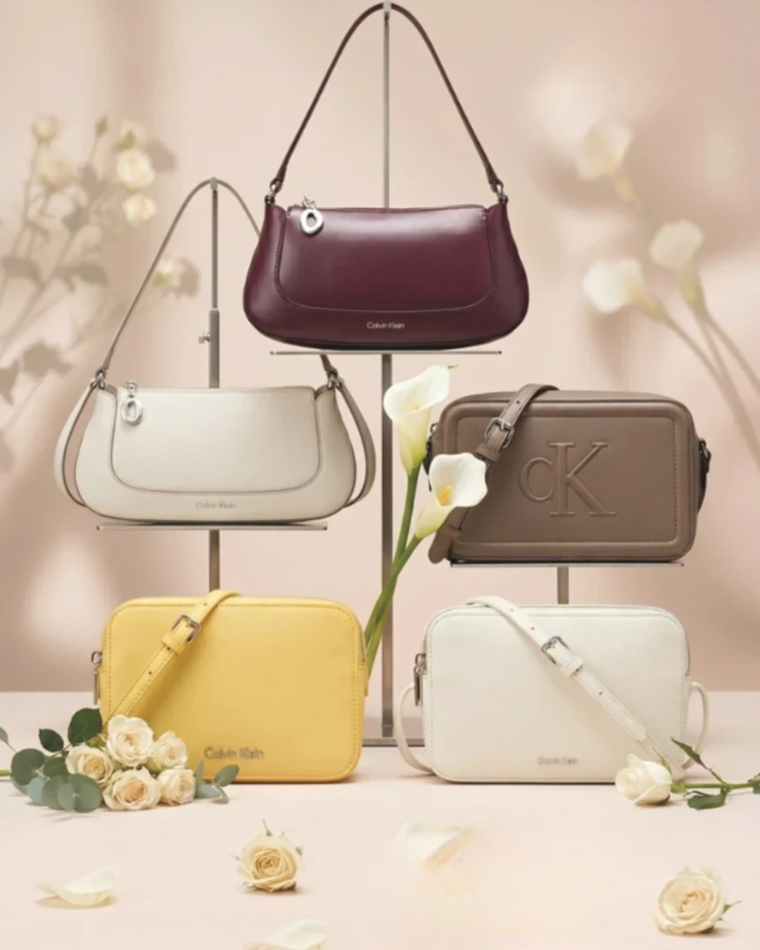 Torebki CK w Kauflandzie od 21.05 Pięć torebek marki Calvin Klein w różnych kolorach — bordowa, biała, żółta, brązowa i kremowa — elegancko wyeksponowane na geometrycznych stojakach, otoczone białymi i kremowymi kwiatami.