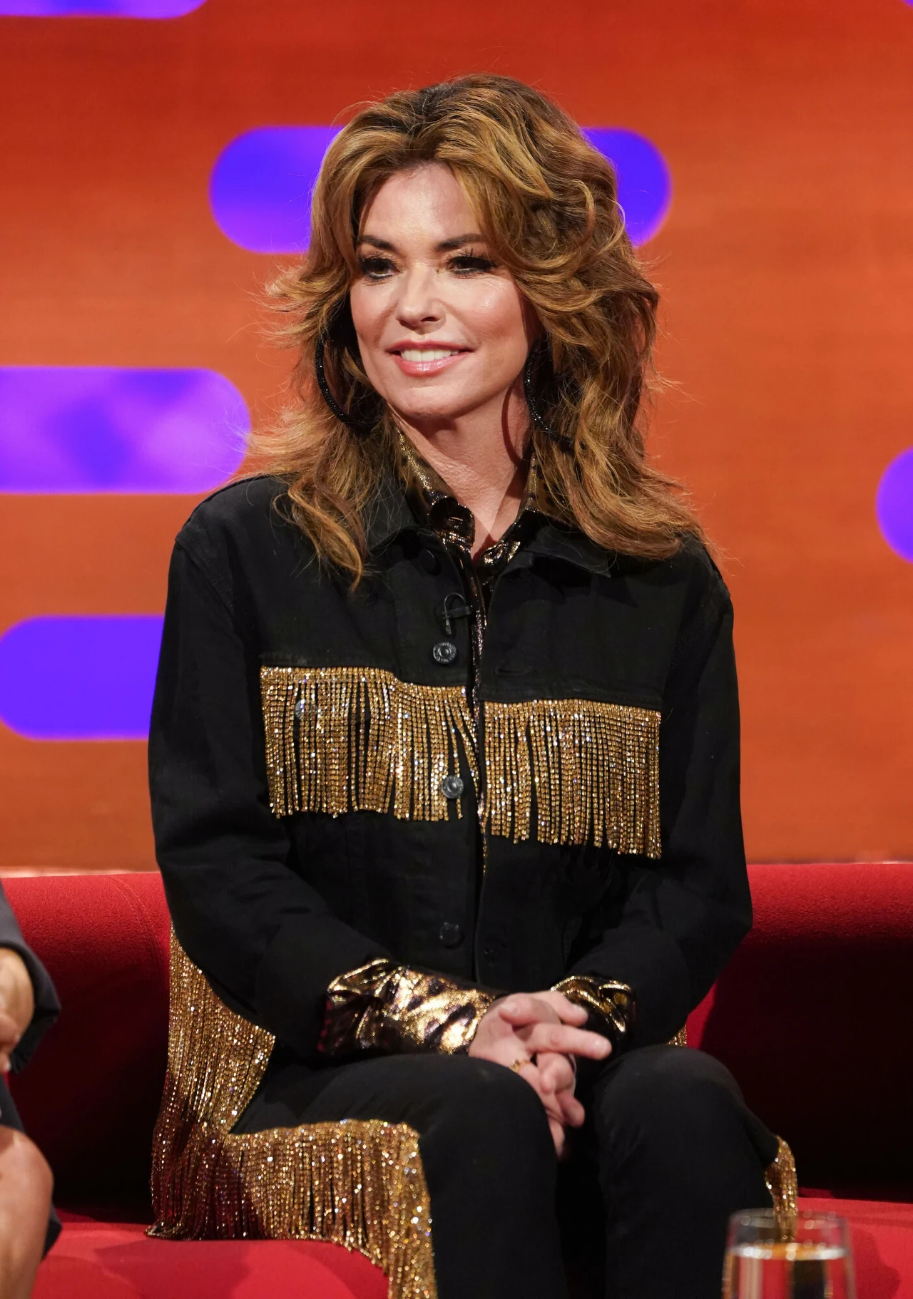Shania Twain w programie Grahama Nortona w 2023 roku Kobieta z burzą brązowych włosów, ubrana w czarną koszulę ozdobioną złotymi frędzlami, siedzi na czerwonej kanapie na tle pomarańczowej ściany z fioletowymi, owalnymi elementami.