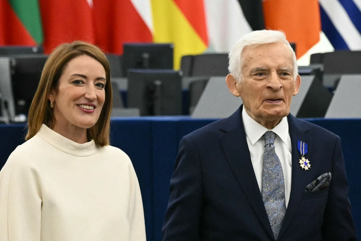 Kobieta w białym ubraniu i starszy mężczyzna w garniturze z odznaczeniem na piersi stoją razem na tle flag państwowych oraz konferencyjnych pulpitów.