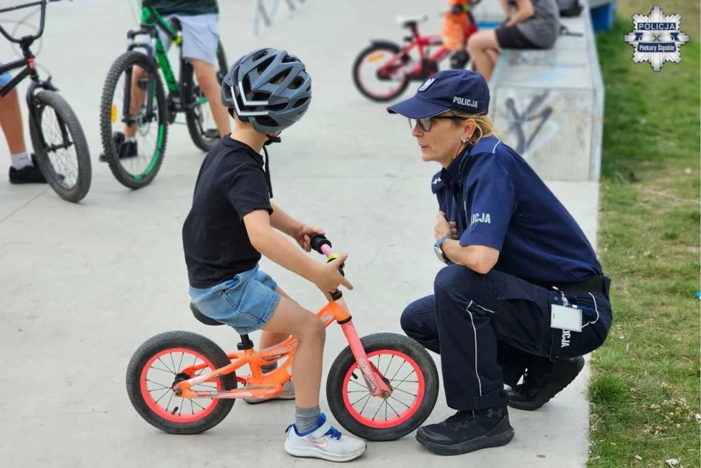 Policjantka w mundurze rozmawia z dzieckiem w kasku siedzącym na rowerze, w tle inni rowerzyści na skateparku.
