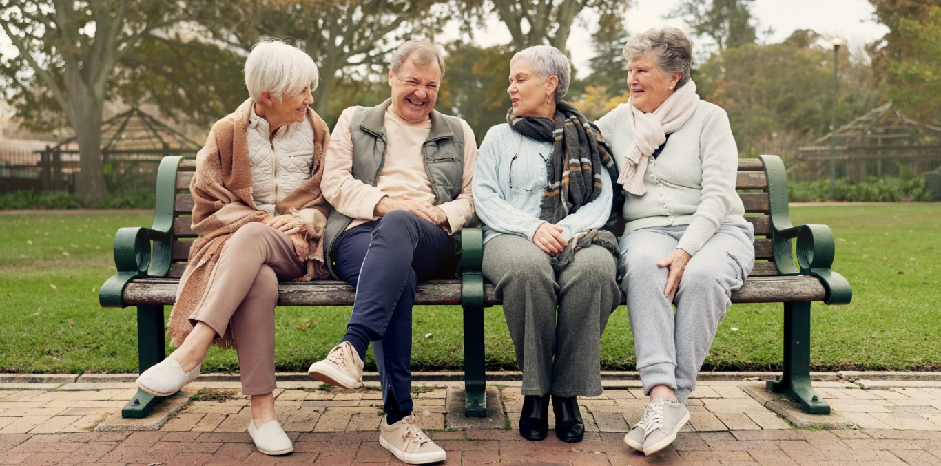 Cztery uśmiechnięte seniorki siedzą obok siebie na ławce w parku, rozmawiając i śmiejąc się podczas wspólnego spotkania.