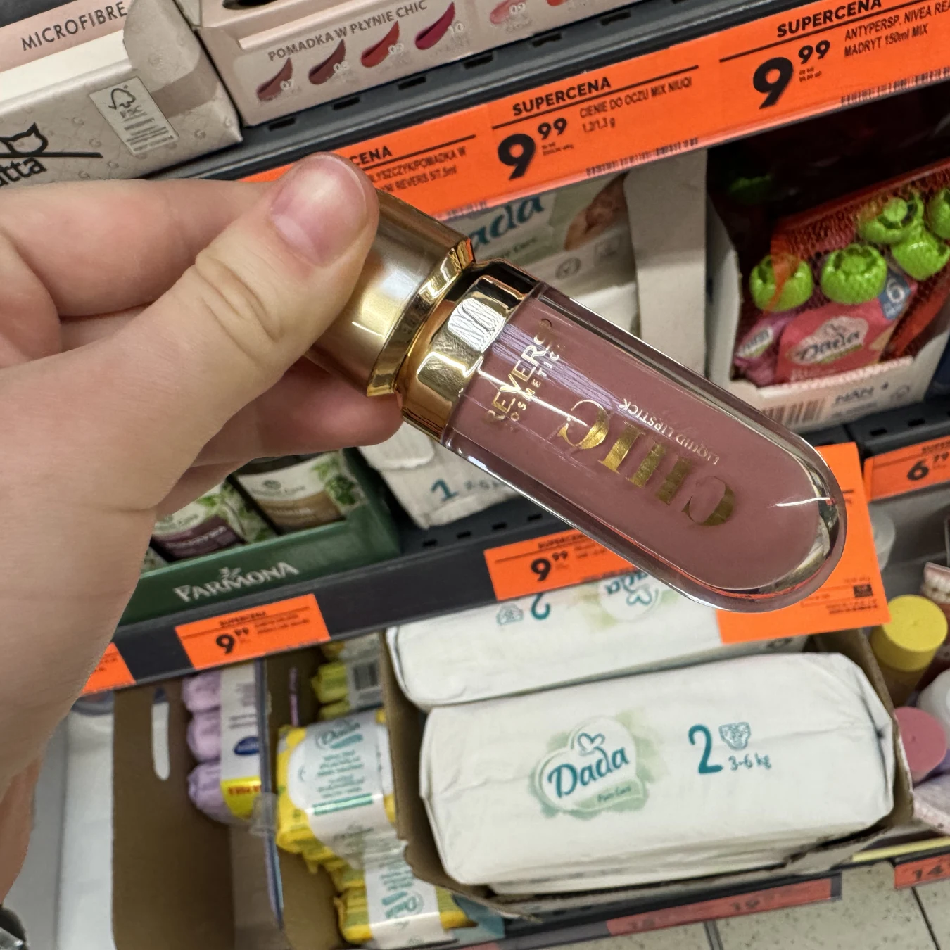 Ręka trzyma szminkę Eveline o złotej nakrętce, w tle półka sklepowa z pieluszkami Dada i innymi produktami oraz pomarańczowymi etykietami cenowymi.