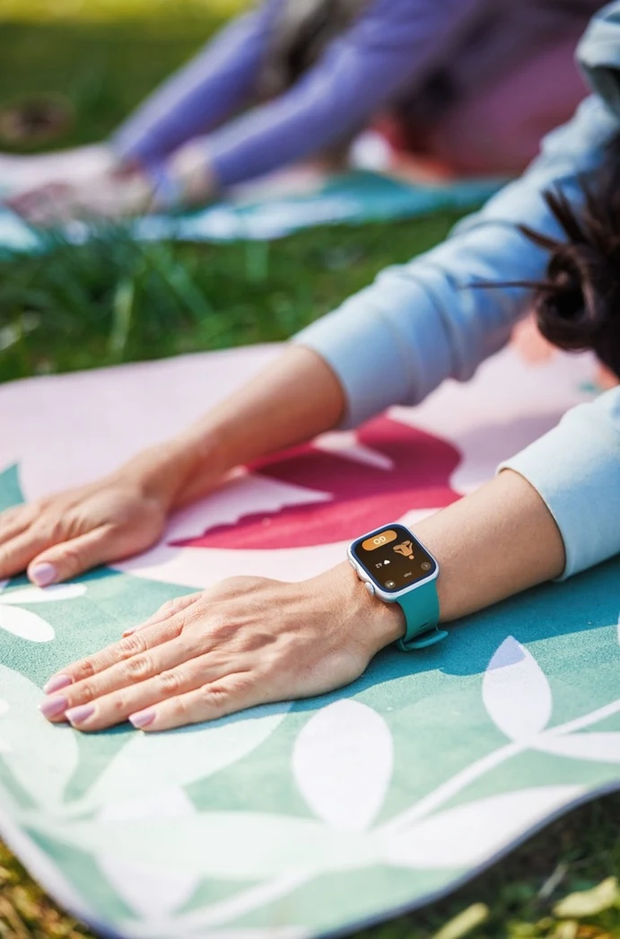 Ręka w niebieskim smartwatchu spoczywająca na kolorowej macie do ćwiczeń na trawie.