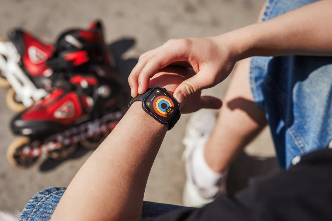Osoba w krótkich spodenkach sprawdzająca smartwatch, obok leżą czerwone rolki.