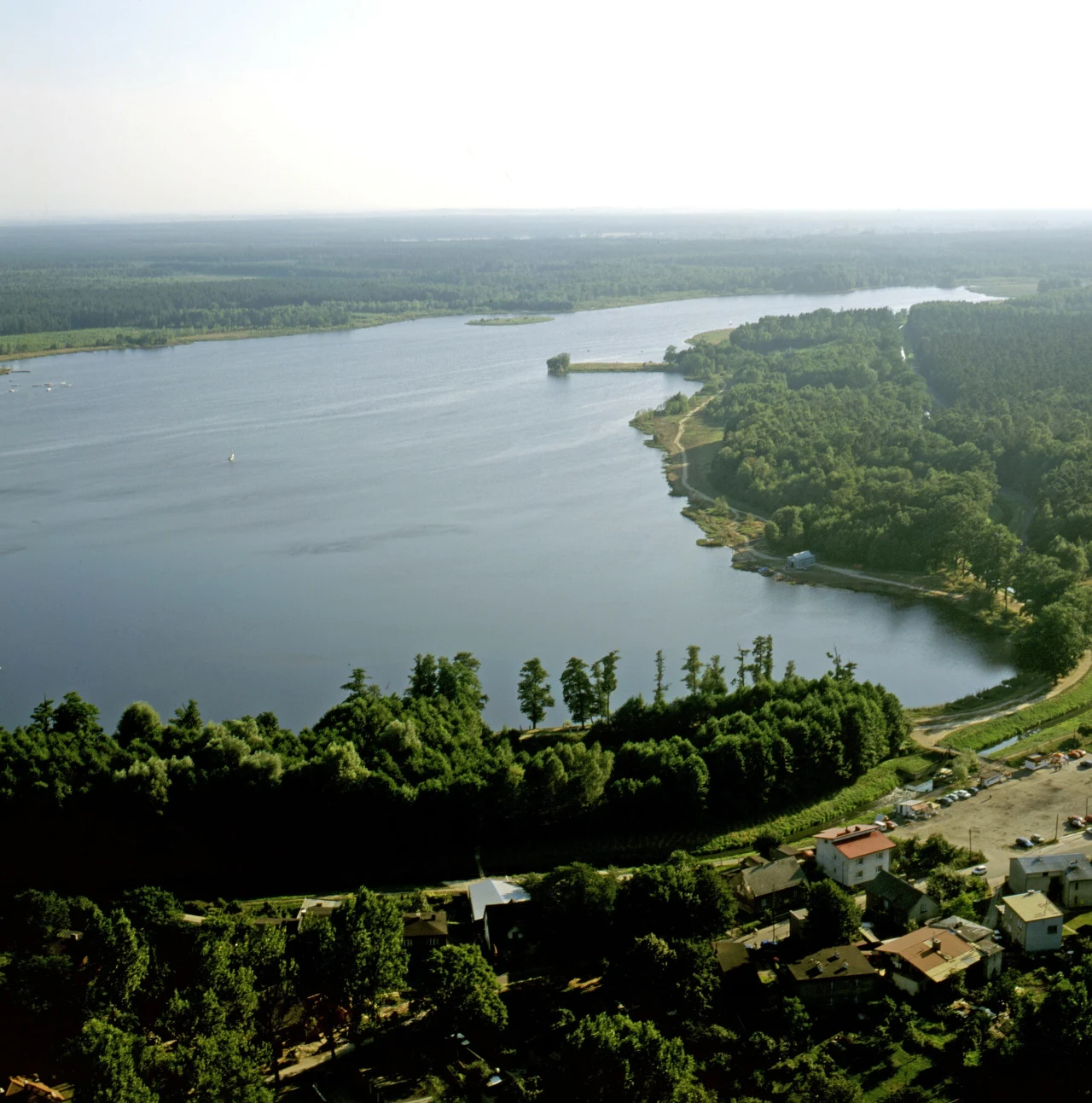 Rozległe jezioro otoczone gęstym lasem, z widoczną zabudową na pierwszym planie oraz naturalną linią brzegową wijącą się wśród zieleni.