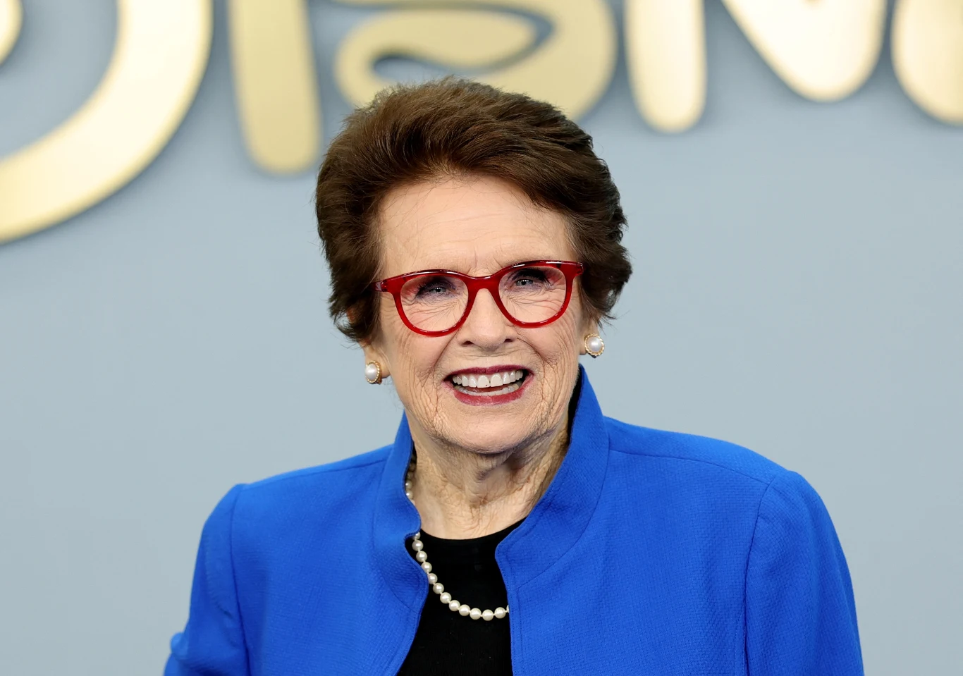 Biellie Jean King Uśmiechnięta kobieta w niebieskiej marynarce, z krótkimi brązowymi włosami, nosząca czerwone okulary i perłowy naszyjnik, stoi na tle jasnego napisu na ścianie.
