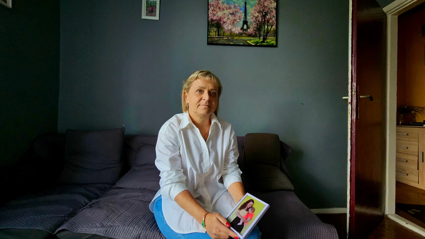 Małgorzata, mama Doroty Kobieta w jasnej koszuli siedzi na kanapie i trzyma w rękach zdjęcie osoby, którą ogląda ze skupieniem; w tle ciemnoniebieska ściana ozdobiona obrazem z wieżą Eiffla.