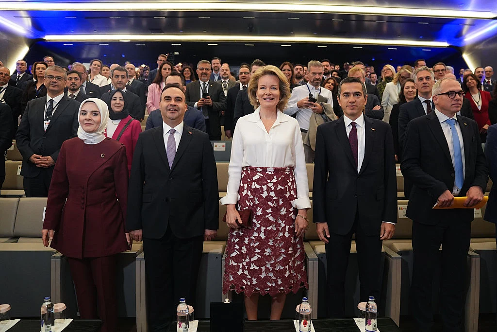 Grupa elegancko ubranych osób stojących w pierwszym rzędzie sali konferencyjnej, wśród których wyróżnia się kobieta w białej bluzce i bordowej spódnicy, a za nimi liczni uczestnicy wydarzenia zajmujący kolejne rzędy siedzeń.