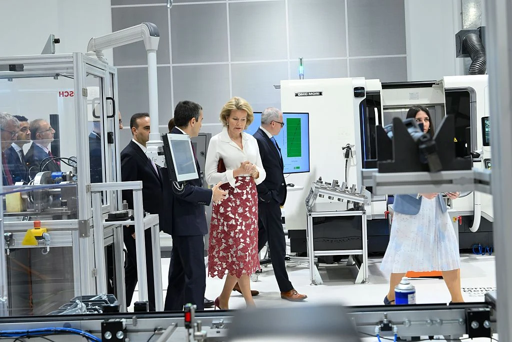 Grupa elegancko ubranych osób spaceruje po nowoczesnej hali produkcyjnej, otoczona zaawansowanymi maszynami przemysłowymi i urządzeniami do obróbki, co sugeruje prezentację lub zwiedzanie infrastruktury technologicznej.