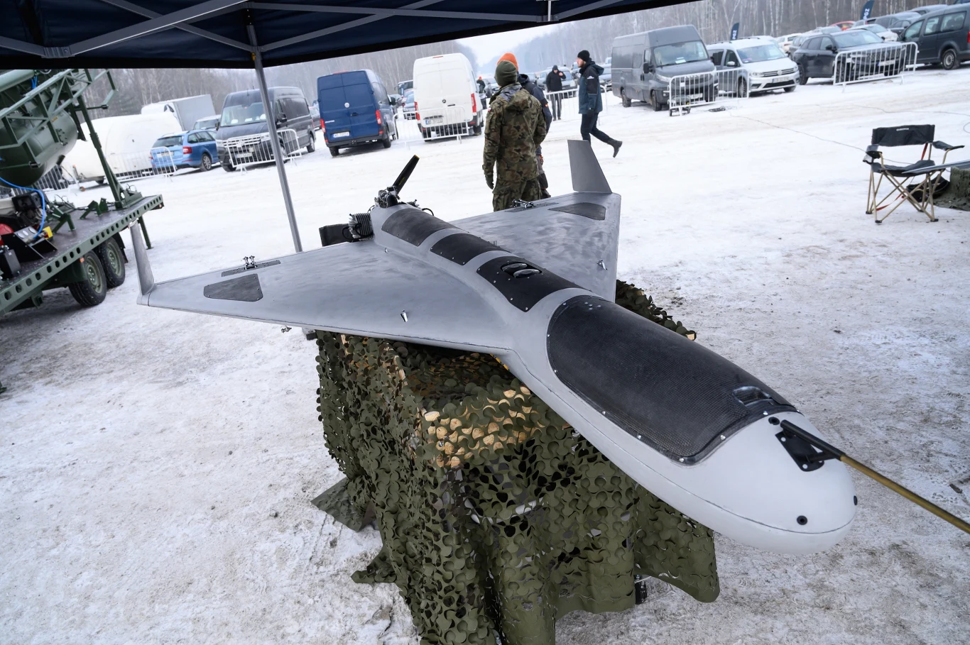 Dron PLArgonia podczas prezentacji na poligonie w Zielonce 19 lutego Duży bezzałogowy dron wojskowy o aerodynamicznym kształcie, ułożony na siatce maskującej, w otoczeniu śnieżnego terenu oraz kilku osób w wojskowych mundurach.
