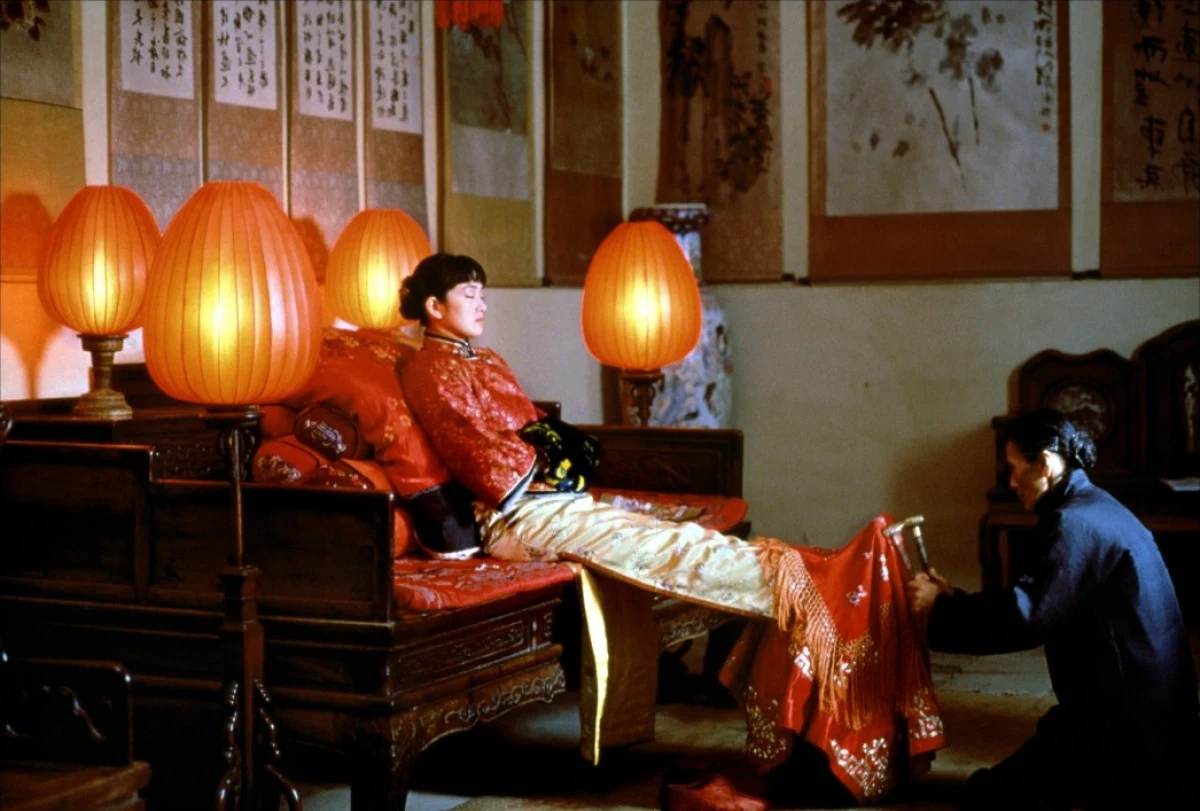 Gong Li w filmie "Zawieście czerwone latarnie" / Gravity Films Kobieta w tradycyjnym, ozdobnym stroju zasiada na drewnianym łóżku otoczonym ciepłym światłem czerwonych lampionów, podczas gdy druga osoba przyklęka u jej stóp i poprawia fragment ubioru.