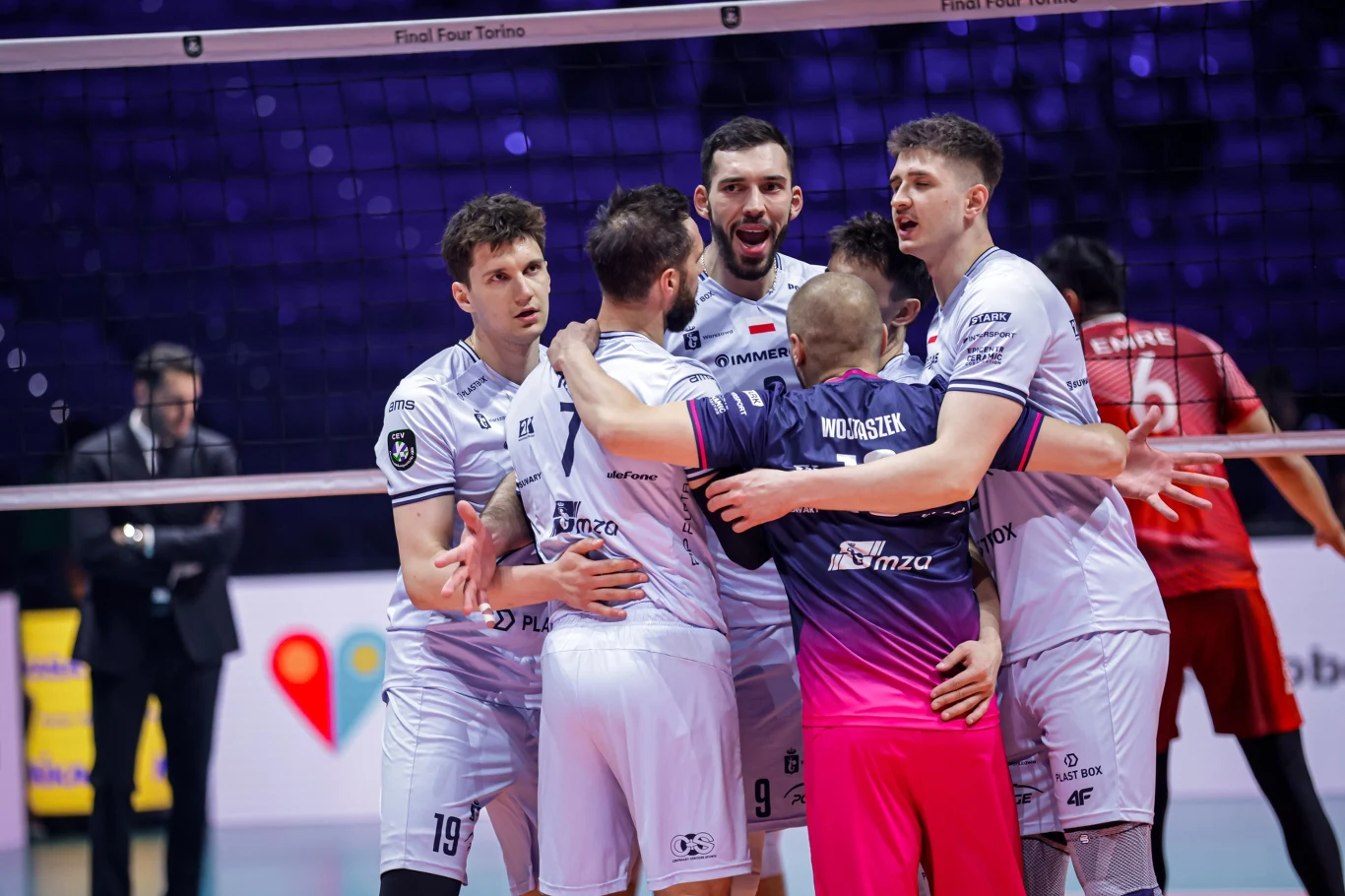 Siatkarze PGE Projektu Warszawa, z prawej Bartosz Gomułka Grupa siatkarzy w białych strojach świętuje zdobyty punkt, przytulając się na środku boiska przed siatką, widoczny jest ich entuzjazm i sportowa radość.
