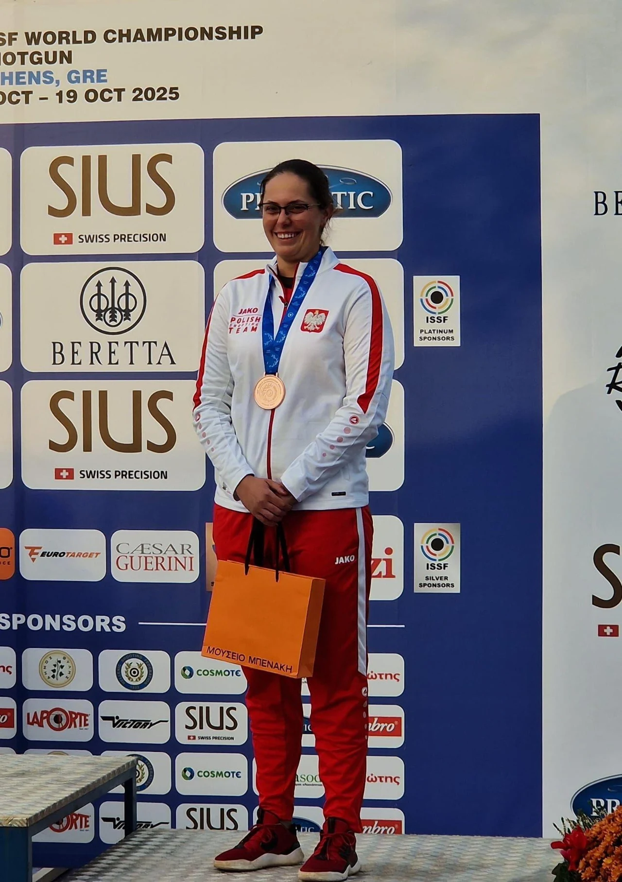 Uśmiechnięta kobieta w biało-czerwonym dresie z medalem na szyi i torbą prezentową stoi na podium przed ścianą z logo sponsorów i organizatorów imprezy sportowej.