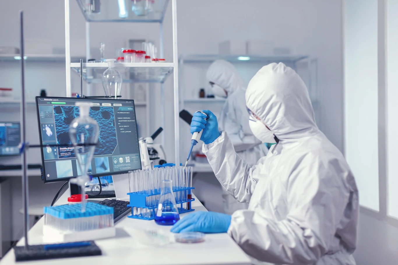 Ebola może być groźna dla życia i zdrowia ludzi Naukowiec w ochronnym kombinezonie analizuje próbki za pomocą pipety, otoczony sprzętem laboratoryjnym i komputerem z wyświetlonymi danymi naukowymi.