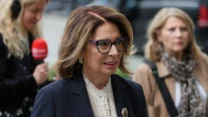 Kobieta w okularach i eleganckim granatowym żakiecie z broszką, ubrana w białą koszulę, idzie ulicą w towarzystwie innych osób.