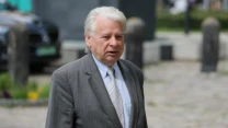 Starszy mężczyzna w szarym garniturze i jasnym krawacie idący po bruku, z poważnym wyrazem twarzy.