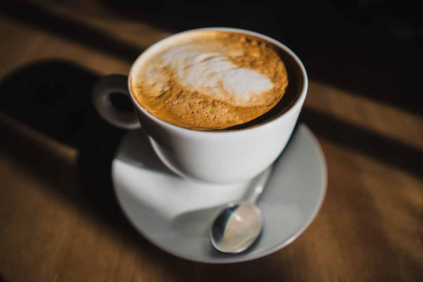 Hojicha to japoński napój pełen wartości, a zarazem bez kofeiny Biała filiżanka napoju hojicha latte z pianką na spodku, obok metalowej łyżeczki, w dziennym świetle.