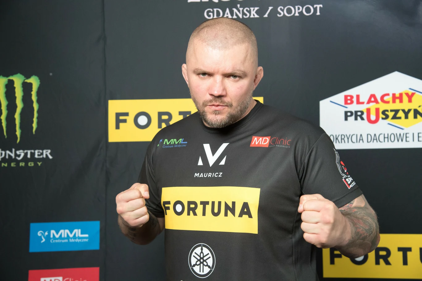 Łukasz "Juras" Jurkowski po raz ostatni w KSW walczył w 2021 roku Mężczyzna z ogoloną głową w czarnej koszulce sportowej z widocznymi logotypami sponsorów pozuje w postawie zawodnika MMA z zaciśniętymi pięściami na tle ściany z reklamami.