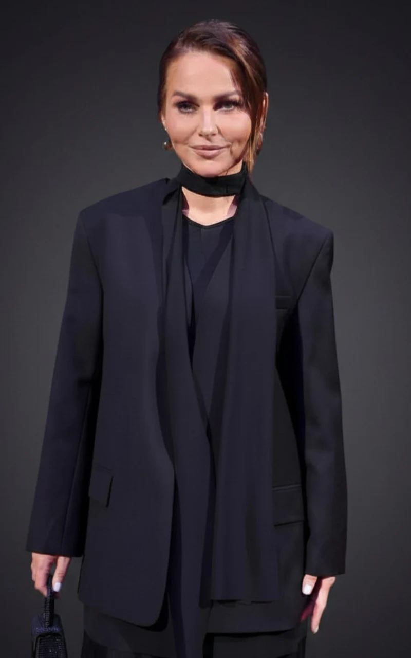 Kobieta w eleganckim czarnym garniturze i apaszce, trzymająca małą torebkę, stoi na jednolitym ciemnym tle.