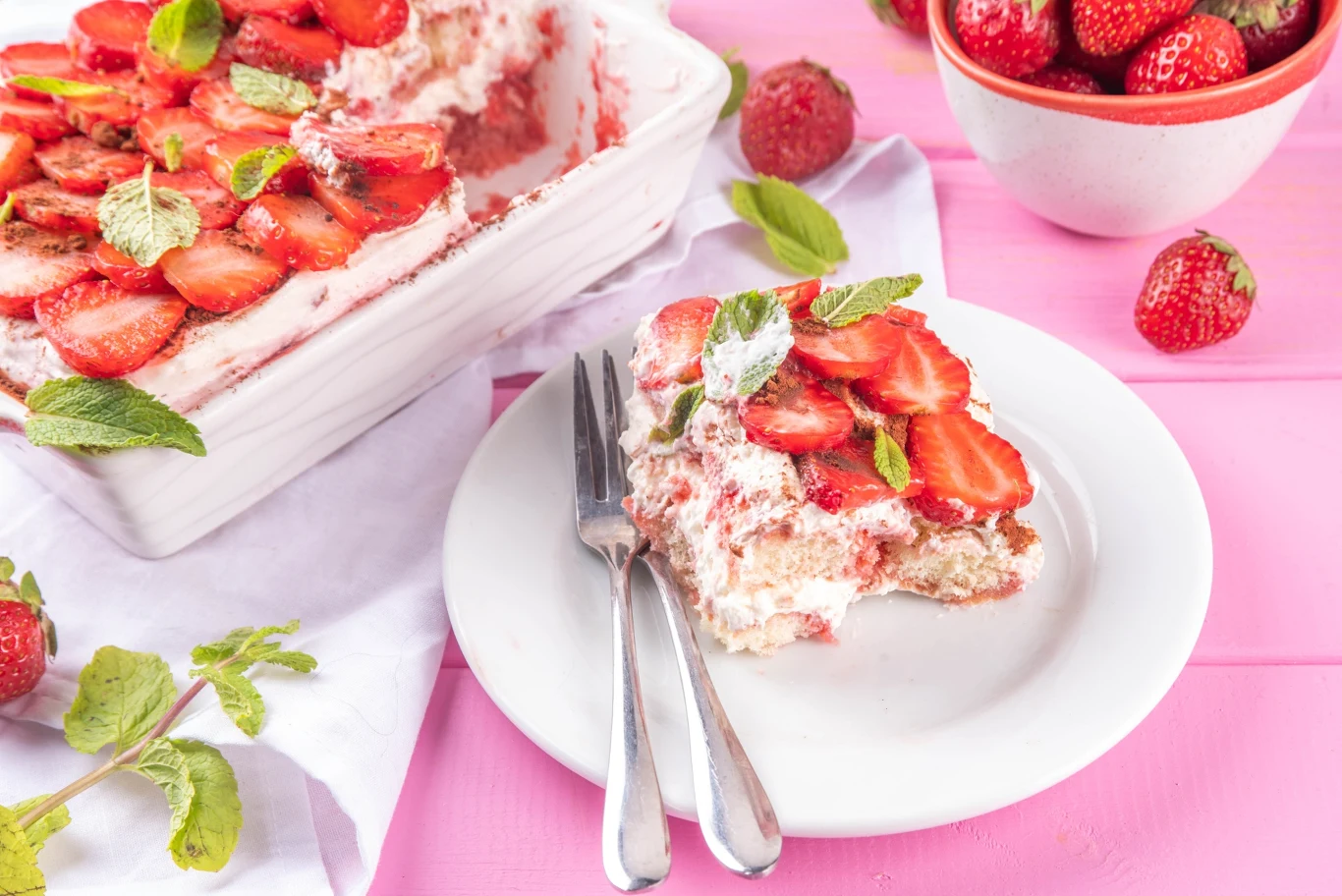 Porcja truskawkowego tiramisu z warstwami kremu i truskawek, udekorowana miętą na białym talerzu, obok sztućców.