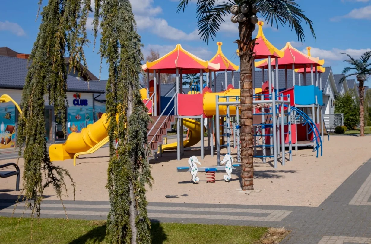 Kolorowy plac zabaw z dużą konstrukcją z czerwonymi i żółtymi daszkami, zjeżdżalnią, wspinaczką oraz huśtawkami sprężynowymi na piaszczystym podłożu, otoczony egzotycznymi drzewami.
