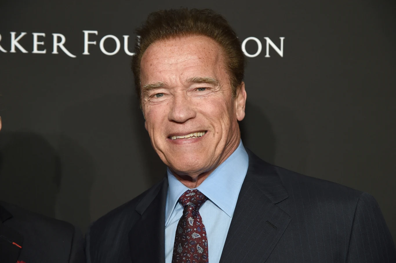 Arnold Schwarzenegger Arnold Schwarzenegger w garniturze i wzorzystym krawacie na tle czarnej ścianki z białym napisem.