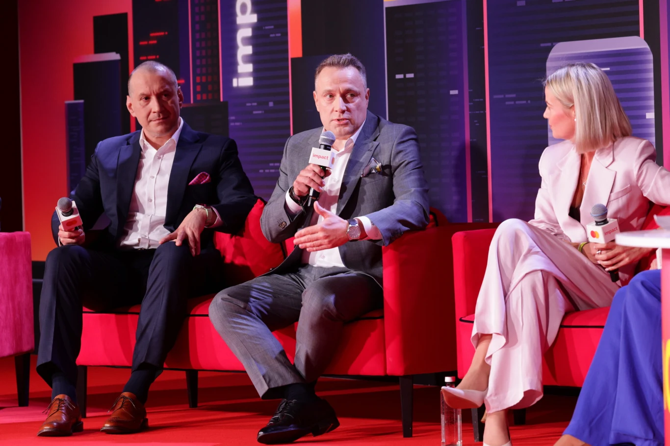 Troje elegancko ubranych osób siedzi na czerwonej sofie, dwie z nich trzymają mikrofony i prowadzą dyskusję na tle stylizowanym na panoramę miejską.