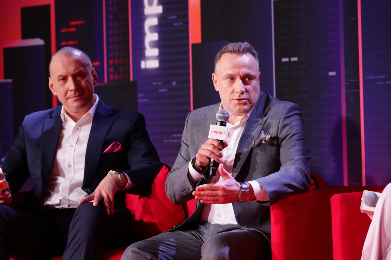 Dwóch elegancko ubranych mężczyzn siedzi na czerwonej sofie podczas panelu dyskusyjnego, jeden z nich trzyma mikrofon i gestykuluje w trakcie wypowiedzi, w tle widoczne elementy scenografii z napisem „impact”.